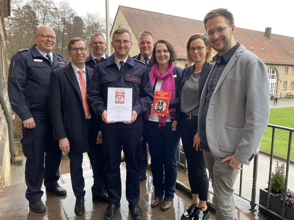 Gruppenfoto von acht Personen, darunter Feuerwehrleute in Uniform und weitere Personen in Business-Kleidung. Eine Person in der Mitte hält eine Urkunde mit Feuerwehr-Logo, eine andere ein rotes Schild mit der Aufschrift ‚Partner der Feuerwehr‘.
