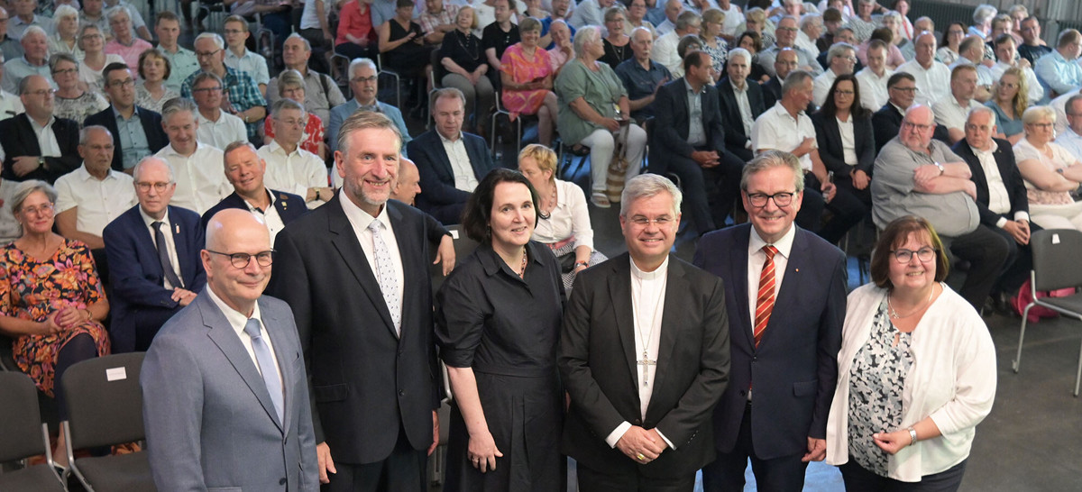 Gruppenfoto mit sechs Personen in der ersten Reihe, darunter Männer im Anzug, eine Frau im schwarzen Kleid und ein Geistlicher mit Kreuzkette. Im Hintergrund sitzt ein großer Saal voller Menschen, die aufmerksam in Richtung Kamera blicken.