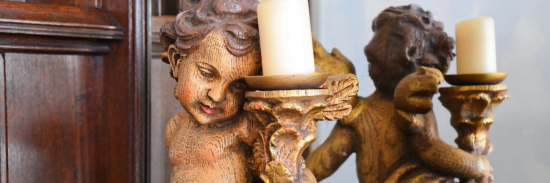 Eine kunstvoll geschnitzte Holzfigur eines Engels oder Putto hält einen goldverzierten Kerzenhalter mit einer weißen Kerze.