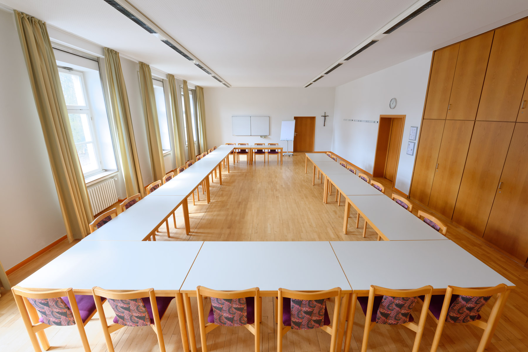 Heller Seminarraum mit U-förmig aufgestellten Tischen und Holzstühlen mit gemusterten Rückenlehnen. Der Raum hat Holzboden, große Fenster mit hellgrünen Vorhängen, eine weiße Wand mit Kreidetafeln und ein Kreuz sowie eine Tür und einen großen Schrank aus Holz.