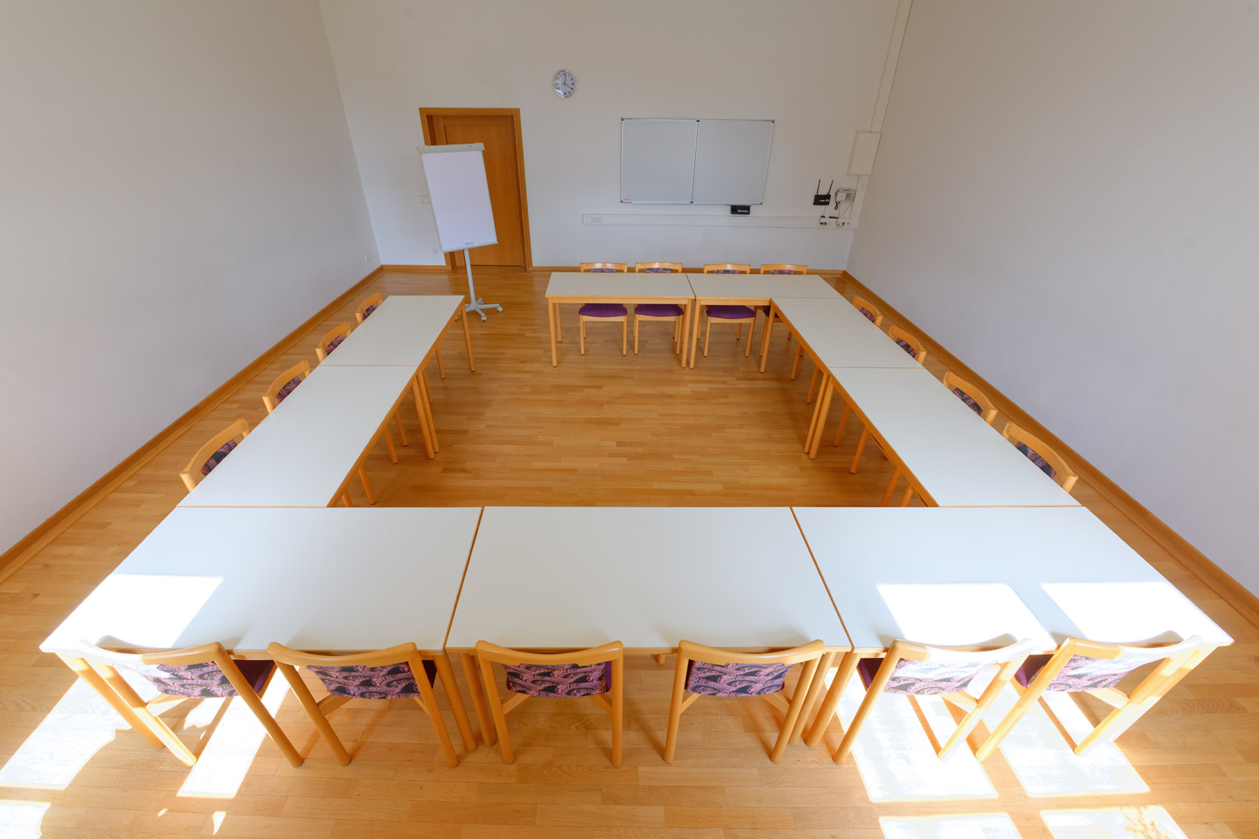 Heller Seminarraum mit U-förmig angeordneten weißen Tischen und Holzstühlen mit gemusterten Rückenlehnen. An der Stirnseite stehen ein Flipchart, eine weiße Tafel und eine Tür aus Holz, der Raum hat Parkettboden und weiße Wände.