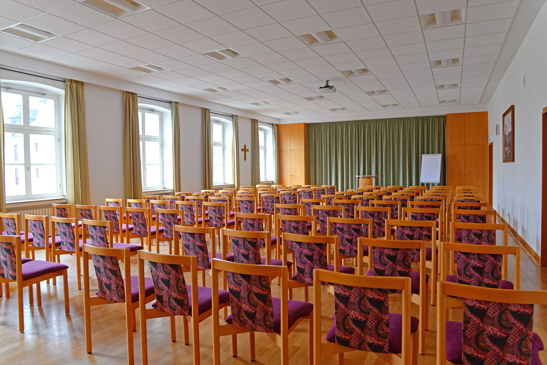 Großer heller Hörsaal mit mehreren Reihen hölzerner Stühle, die violette Sitzflächen und gemusterte Rückenlehnen haben. Der Raum hat Holzboden, grüne Vorhänge, große Fenster an der linken Seite und eine Bühne mit Rednerpult vorne.