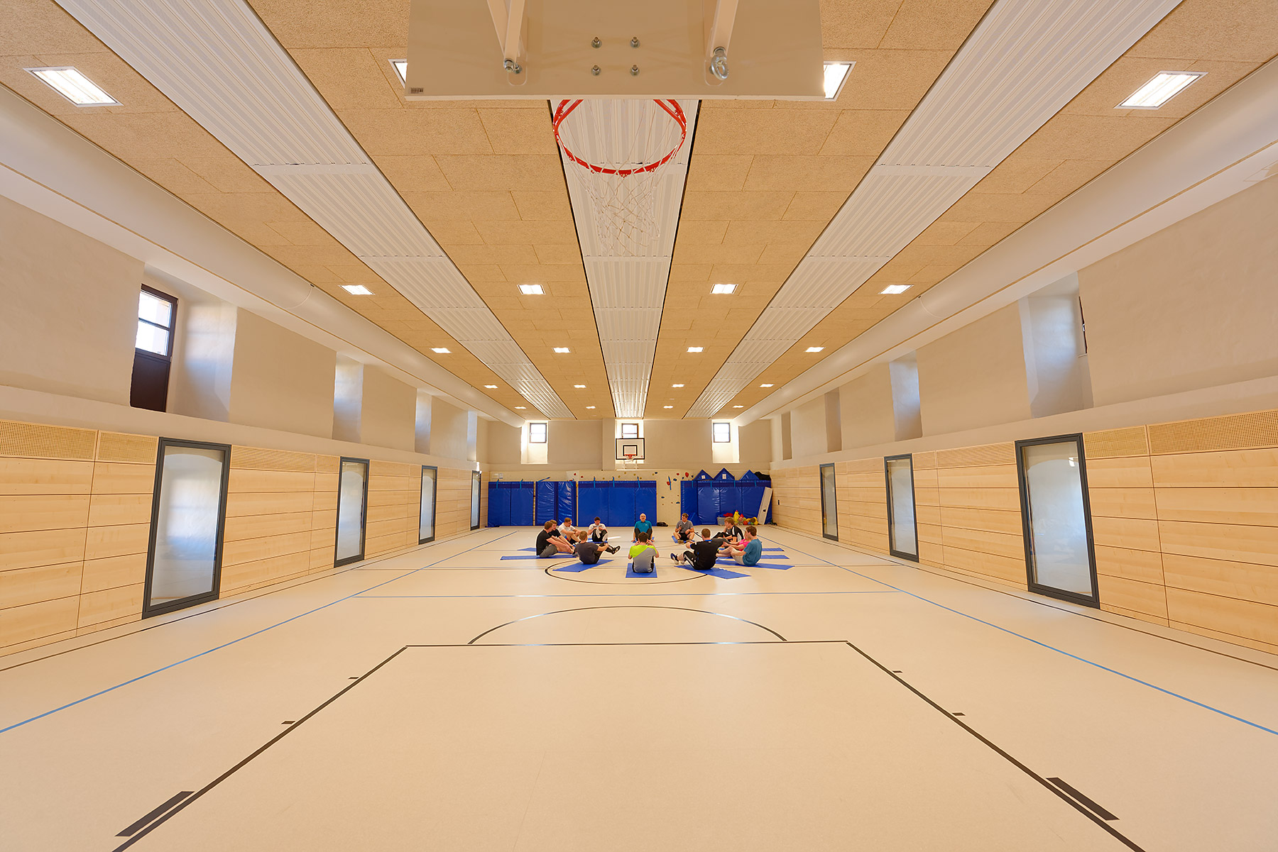 Helle Turnhalle mit Holzwänden und Basketballkörben, in der eine Gruppe von Personen auf Matten im Kreis sitzt.