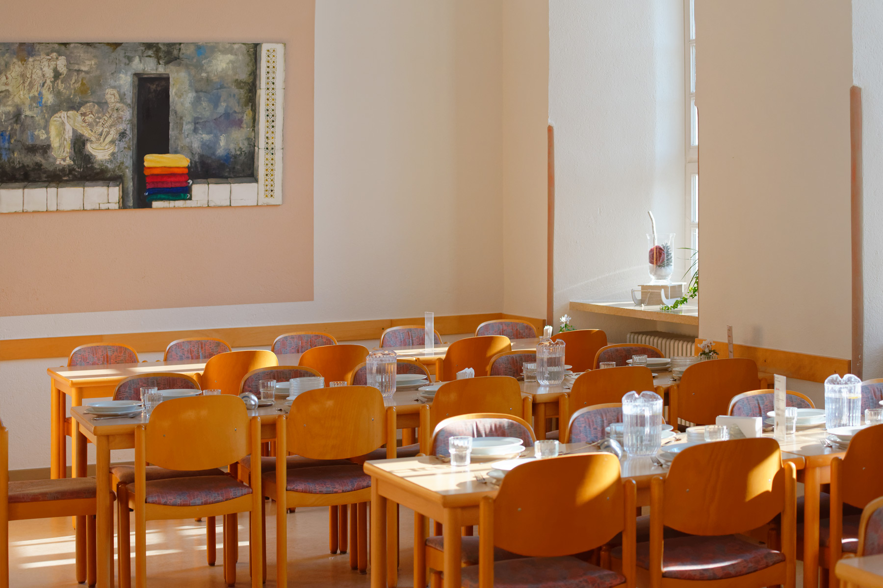 Heller Speisesaal mit mehreren gedeckten Holztischen und Stühlen, Krügen und Gläsern. An der Wand hängt ein farbenfrohes Gemälde, und durch das Fenster fällt Sonnenlicht in den Raum.
