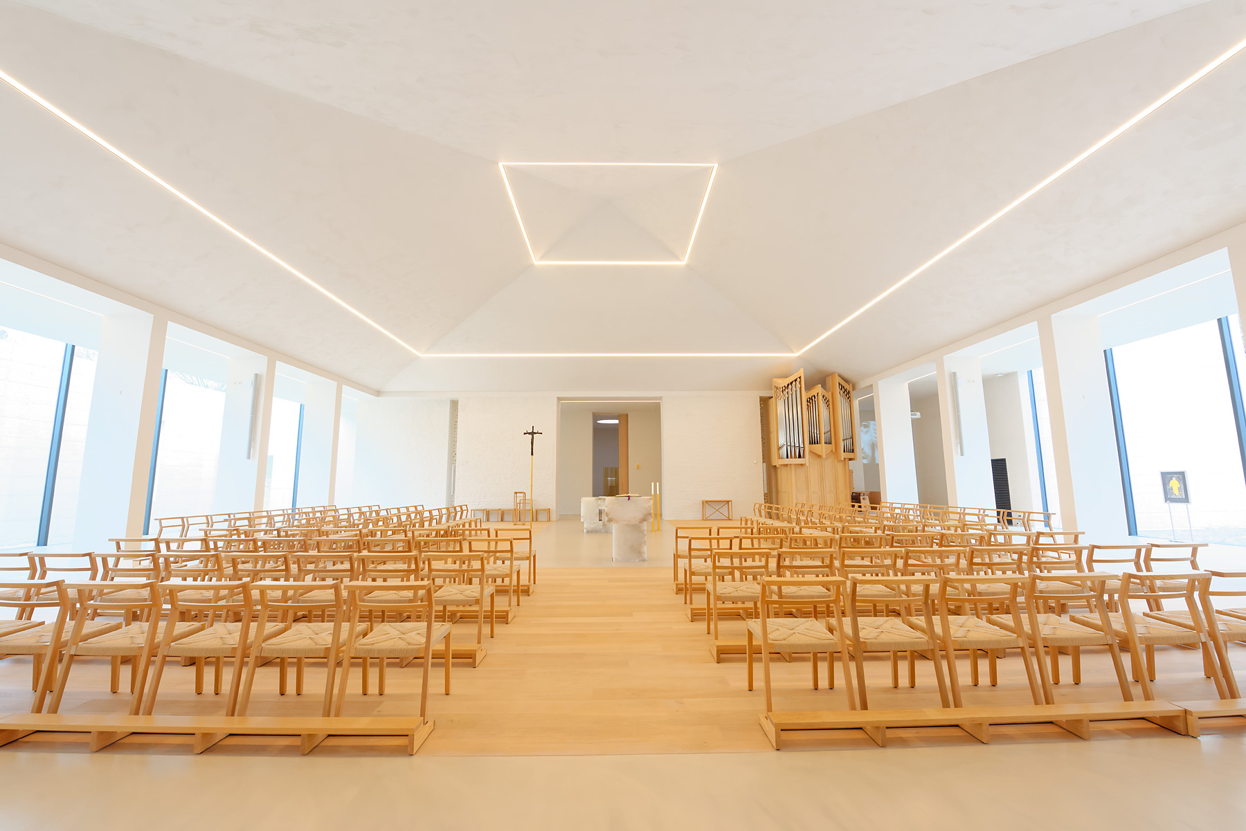 Heller, moderner Kirchenraum mit schlichten Holzbänken, zentralem Altar und klarer geometrischer Deckenbeleuchtung. Durch große Fenster fällt natürliches Licht in den Raum.