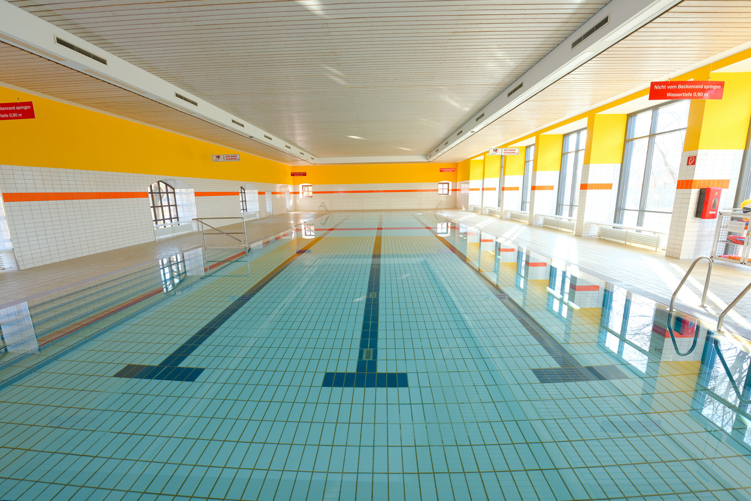 Helles Hallenbad mit einem großen Schwimmbecken, blauer Kachelboden mit Bahnenmarkierungen, gelb-weißen Wänden und großen Fenstern, die Tageslicht hereinlassen.