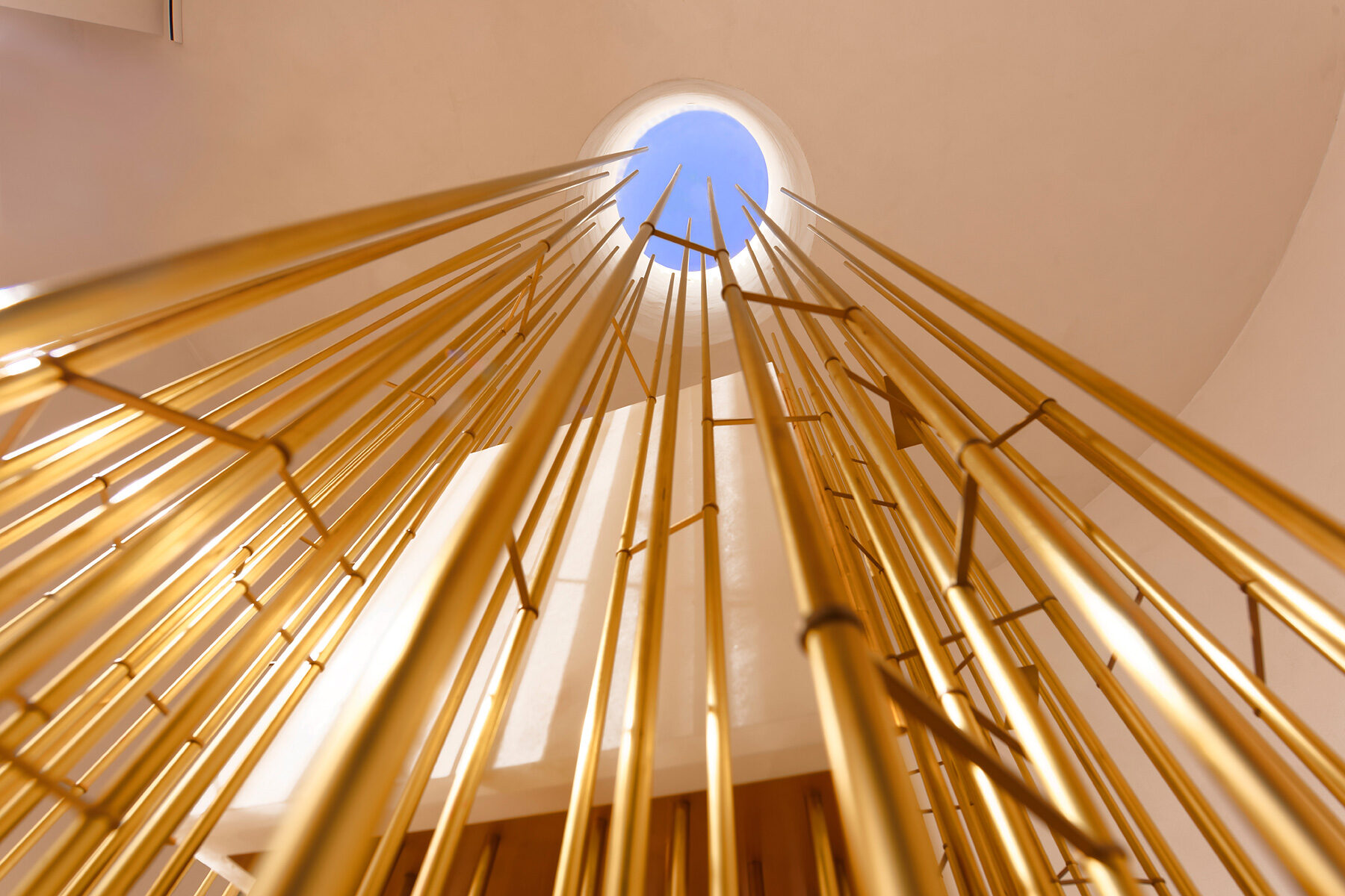 Blick von unten auf eine Installation aus vielen goldfarbenen Stäben, die sich nach oben zu einer runden Öffnung in der Decke hin ausrichten, durch die blaues Himmelslicht einfällt.
