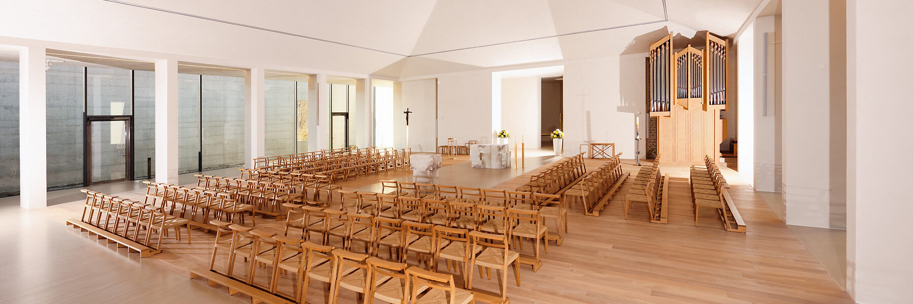 Innenansicht einer modernen, hellen Kirche mit hellem Holzfußboden und weißen Wänden. In der Mitte stehen Holzbänke und Stühle in rechteckiger Anordnung um einen schlichten Altar aus Stein. Rechts befindet sich eine große Orgel.