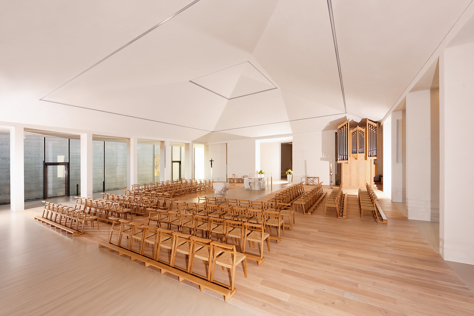 Innenansicht einer modernen, hellen Kirche mit hellem Holzfußboden und weißen Wänden. In der Mitte stehen Holzbänke und Stühle in rechteckiger Anordnung um einen schlichten Altar aus Stein. Rechts befindet sich eine große Orgel.