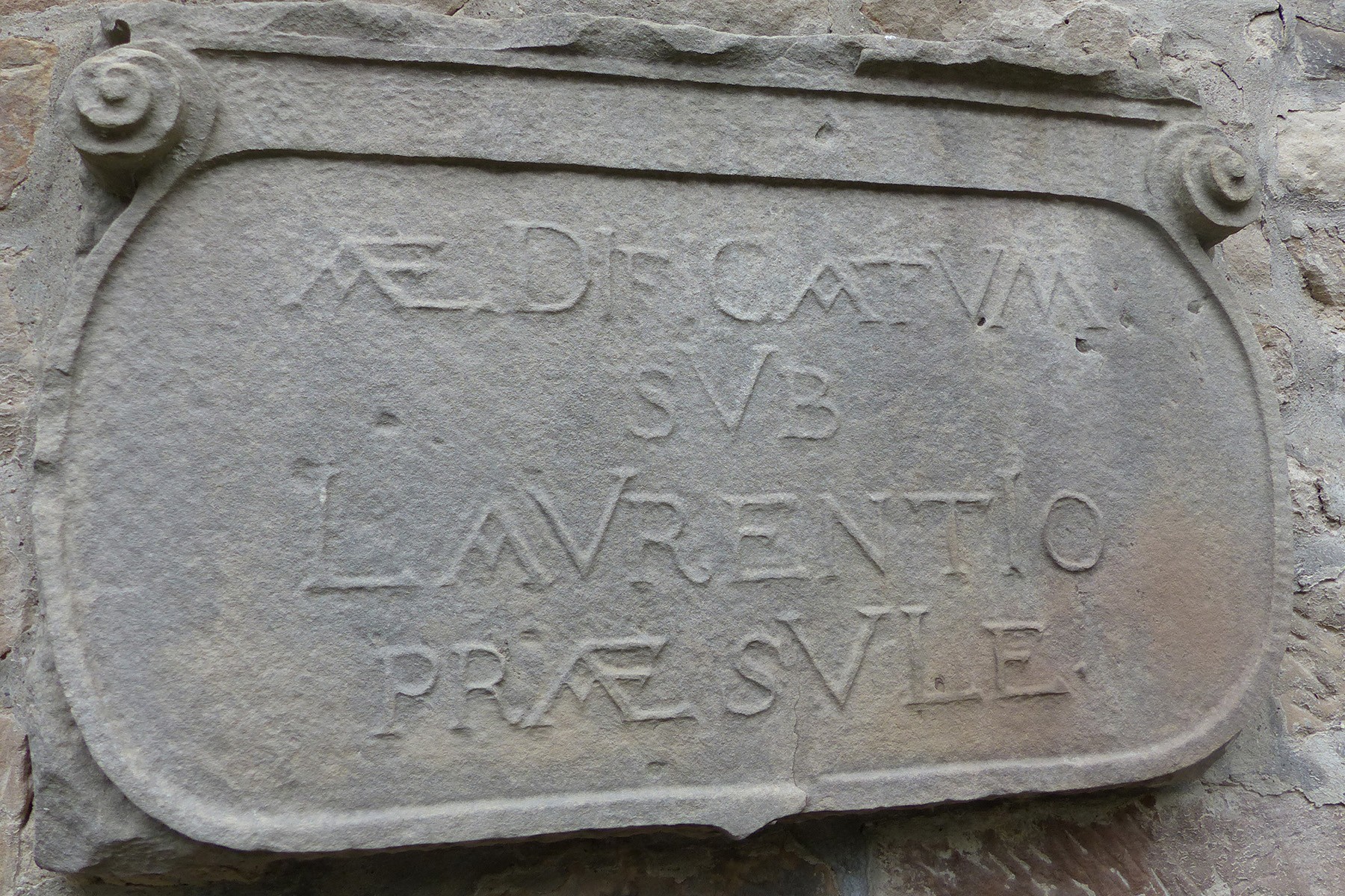 Eine in Stein gehauene Inschrift in lateinischer Sprache ist in eine rechteckige, verzierte Steinplatte mit eingerollten Ecken eingraviert.