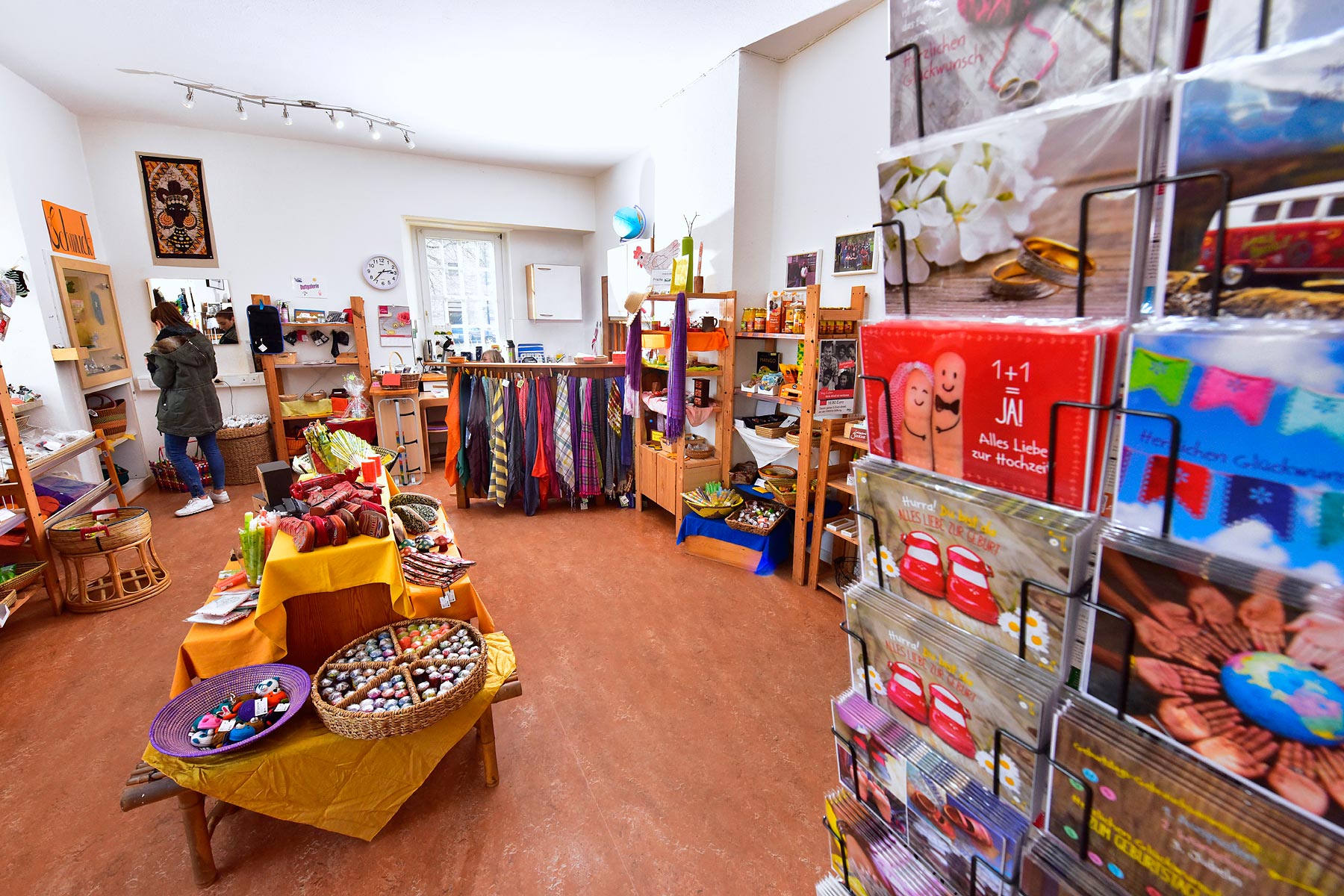 Bunter Weltladen mit Regalen voller fair gehandelter Waren, Kartenständern, Tüchern und Geschenkartikeln. Eine Person schaut sich Produkte an der Wand an.