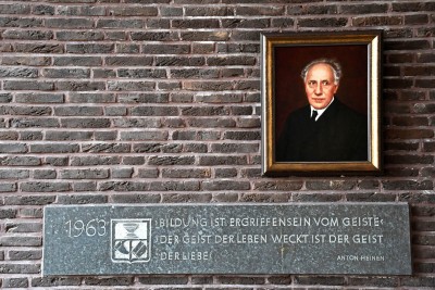 Ein gemaltes Porträt eines Mannes in dunkler Kleidung hängt an einer Backsteinwand. Darunter befindet sich eine Gedenktafel mit der Inschrift „1963 – Bildung ist Ergriffensein vom Geiste; der Geist, der Leben weckt, ist der Geist der Liebe. – Anton Heinen“.