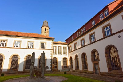 Blick in einen sonnigen Innenhof eines historischen Gebäudes mit hellen Fassaden, großen Rundbogenfenstern und rotem Ziegeldach.