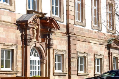 Ein barockes Gebäude mit rot-beiger Sandsteinfassade und weißen Fenstern. Im Mittelpunkt steht ein reich verzierter Eingangsbereich mit Säulen, Rundbogen und Giebel über einer weißen Tür.