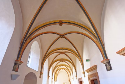 Ein heller Klostergang mit hohen, spitzbogigen Gewölben und farbig gefassten Rippen in Gelb, Rot und Blau.