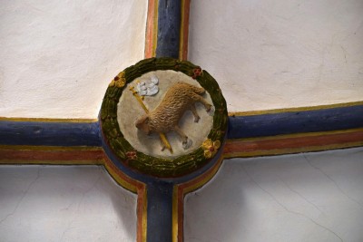 Ein farbig gestaltetes Schlusssteinrelief an einer Gewölbedecke zeigt ein Lamm mit einer Fahne, umgeben von einem grünen, blumenverzierten Kranz.