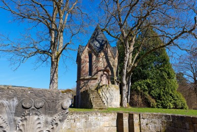Eine kleine historische Kapelle aus Sandstein mit spitzem Schieferdach steht zwischen zwei großen, kahlen Bäumen.
