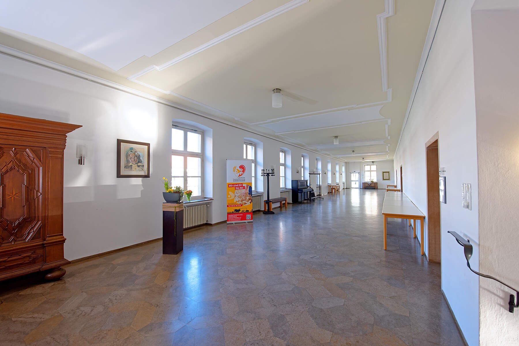 Heller, langer Flur mit großen Fenstern, hellem Boden und dekorativem Stuck an der Decke. Entlang der Wände stehen Tische, ein Klavier, Garderobenständer und ein großer Holzschrank.