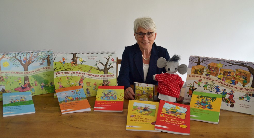 Eine ältere Frau mit kurzen grauen Haaren und Brille sitzt an einem Tisch und lächelt in die Kamera. Vor ihr liegen mehrere farbenfrohe Kinderbücher der Reihe „Klara und die Mäuse“, daneben steht eine graue Maus-Handpuppe im roten Kleid. Im Hintergrund sind weitere illustrierte Buchaufsteller zu sehen.