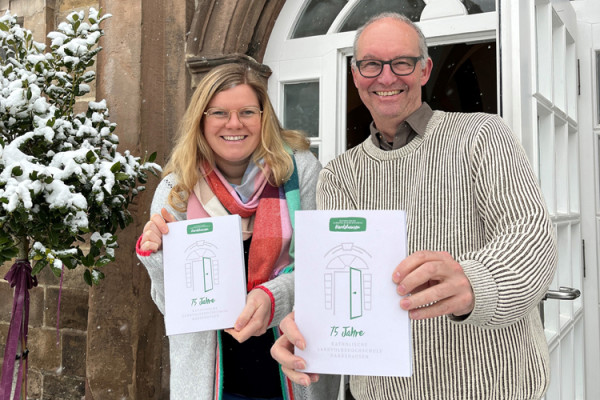 Zwei Personen stehen vor einem Gebäude mit einem weißen Eingang und einer schneebedeckten Umgebung. Beide halten jeweils ein Heft mit einer stilisierten Türgrafik und der Aufschrift „75 Jahre“ auf dem Titel. Eine der Personen trägt einen gestreiften Pullover, die andere einen farbigen Schal. Links im Bild steht ein ebenfalls verschneiter kleiner Baum.