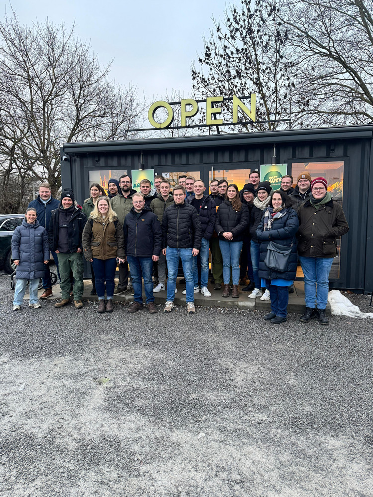 Eine größere Gruppe von Menschen steht draußen vor einem modernen schwarzen Containergebäude mit großen Fenstern. Über dem Eingang hängt ein großes Schild mit der Aufschrift „OPEN“. Die Personen tragen winterliche Kleidung, und auf dem Boden liegt etwas Schnee. Im Hintergrund sind kahle Bäume zu sehen.