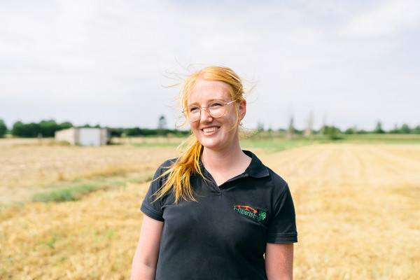 Eine Person mit blondem, im Wind wehenden Haar steht auf einem weitläufigen, goldgelben Feld. Sie trägt ein schwarzes Poloshirt mit einem farbigen Logo auf der linken Brust. Im Hintergrund sind offene Ackerflächen, einige niedrige Gebäude und eine Baumreihe unter einem leicht bewölkten Himmel zu sehen.