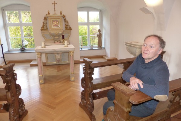 Eine Person sitzt in einer kleinen, hellen Kapelle auf einer kunstvoll geschnitzten Holzbank. Vor der Person steht ein heller Altar mit goldenen Verzierungen, einem Kreuz und einem Kelch. Durch zwei große Fenster hinter dem Altar fällt Tageslicht in den Raum. Rechts neben dem Altar steht eine Statue, links ist ein Taufbecken zu sehen.