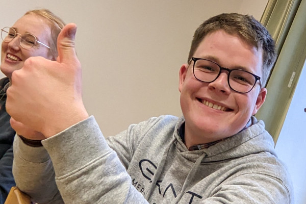 Eine Person sitzt an einem Tisch und zeigt mit der rechten Hand demonstrativ den Daumen nach oben. Sie trägt einen hellgrauen Kapuzenpullover mit dunkelblauem Schriftzug. Neben ihr sitzt eine weitere Person, deren Kopf teilweise im Bild erscheint. Im Hintergrund sind eine beige Wand und ein Fenster mit hellem Vorhang zu sehen.