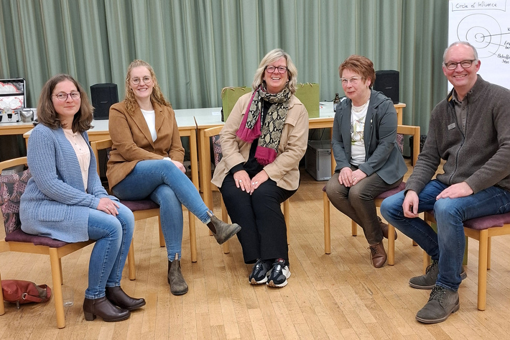 Fünf Personen sitzen in einem Halbkreis auf Stühlen in einem Raum mit Holzfußboden und einem grünen Vorhang im Hintergrund. Vor ihnen stehen mehrere Tische. Rechts ist ein Flipchart mit einer gezeichneten Grafik zu sehen. Die Personen tragen überwiegend legere oder geschäftlich-legere Kleidung.