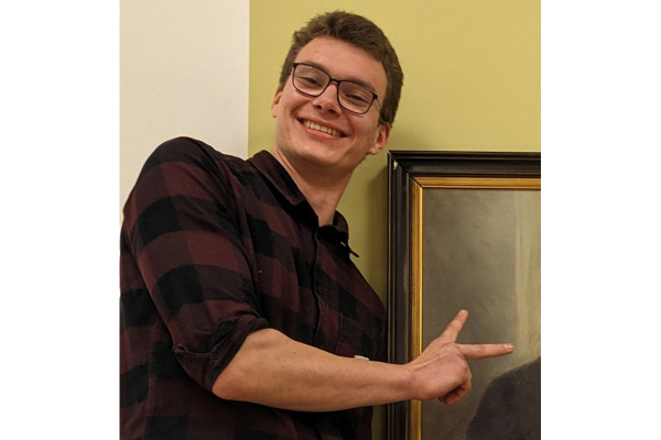 Eine Person steht vor einer zweifarbigen Wand und lehnt sich leicht nach links. Sie trägt ein dunkel kariertes Hemd und zeigt mit der rechten Hand mit zwei ausgestreckten Fingern auf den Rand eines gerahmten Gemäldes, das neben ihr hängt.