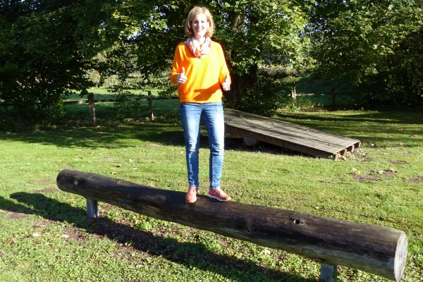 Eine Person steht auf einem liegenden, runden Holzstamm in einem grünen Außenbereich. Die Person trägt ein leuchtend oranges Oberteil, eine Jeans und rote Schuhe und zeigt mit beiden Händen einen Daumen-hoch. Im Hintergrund befinden sich Bäume und eine schräge hölzerne Plattform.