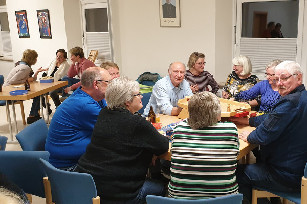 Eine Gruppe von Menschen sitzt in einem hellen Gemeinschaftsraum an mehreren Tischen und spielt gemeinsam ein Brettspiel. Getränke stehen auf den Tischen, und die Personen unterhalten sich angeregt. Im Hintergrund sind weitere Spielrunden und einige Wandbilder zu sehen.