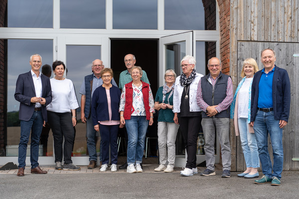 Eine Gruppe von Menschen steht vor einem Gebäude mit großen Glasfenstern und einer geöffneten Tür. Die Personen stehen locker nebeneinander und blicken in Richtung Kamera. Sie tragen überwiegend Freizeit- oder Alltagskleidung, darunter Jacken, Westen und Schals. Die Szene wirkt wie ein Gruppenfoto bei einer Veranstaltung oder einem Treffen im Freien.