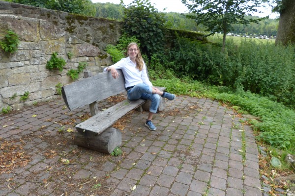 Eine Person sitzt entspannt auf einer schlichten Holzbank auf einem gepflasterten Platz im Freien. Die Person trägt ein helles Oberteil, eine knielange Hose und dunkle Schuhe. Hinter der Bank steht eine niedrige Natursteinmauer, und ringsum wachsen dichte grüne Pflanzen und Bäume. In der Ferne ist eine Wiese zu erkennen.