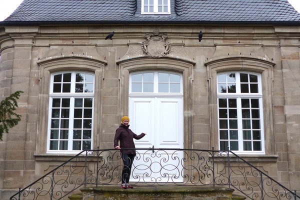 Eine Person steht auf einer kleinen Steintreppe mit verziertem Metallgeländer vor einem historischen Gebäude mit drei großen weißen Doppelfenstern und einer weißen Eingangstür. Die Person trägt dunkle Kleidung und eine gelbe Mütze und zeigt mit einer Hand auf die Tür. Über der Tür befindet sich ein ornamentales Wappen.
