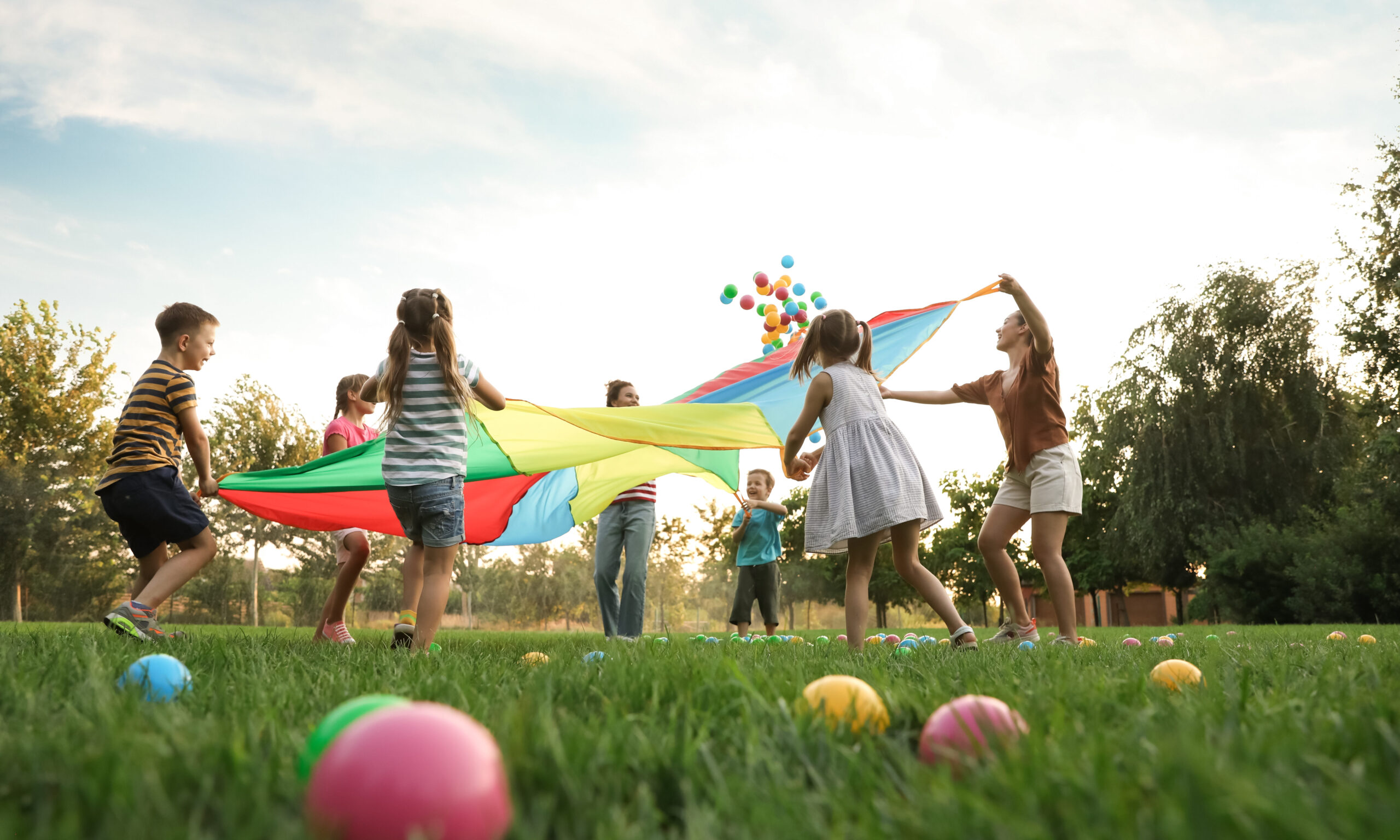 Kinder und pädagogische Fachkräfte spielen gemeinsam mit einem bunten Schwungtuch auf einer Wiese im Freien. Die Gruppe steht im Kreis, während bunte Bälle durch die Luft fliegen. Die Szene vermittelt Freude, Bewegung, Gemeinschaft und spielerisches Lernen in der Kita.